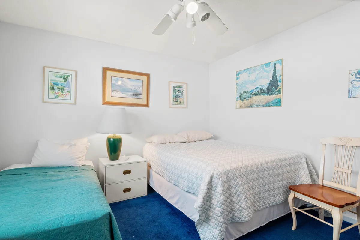 A calm, sunlit bedroom with two beds dressed in blue bedspreads, wall artwork, and a lamp. The atmosphere is serene and tidy, ideal for families or friends needing flexible sleeping arrangements.