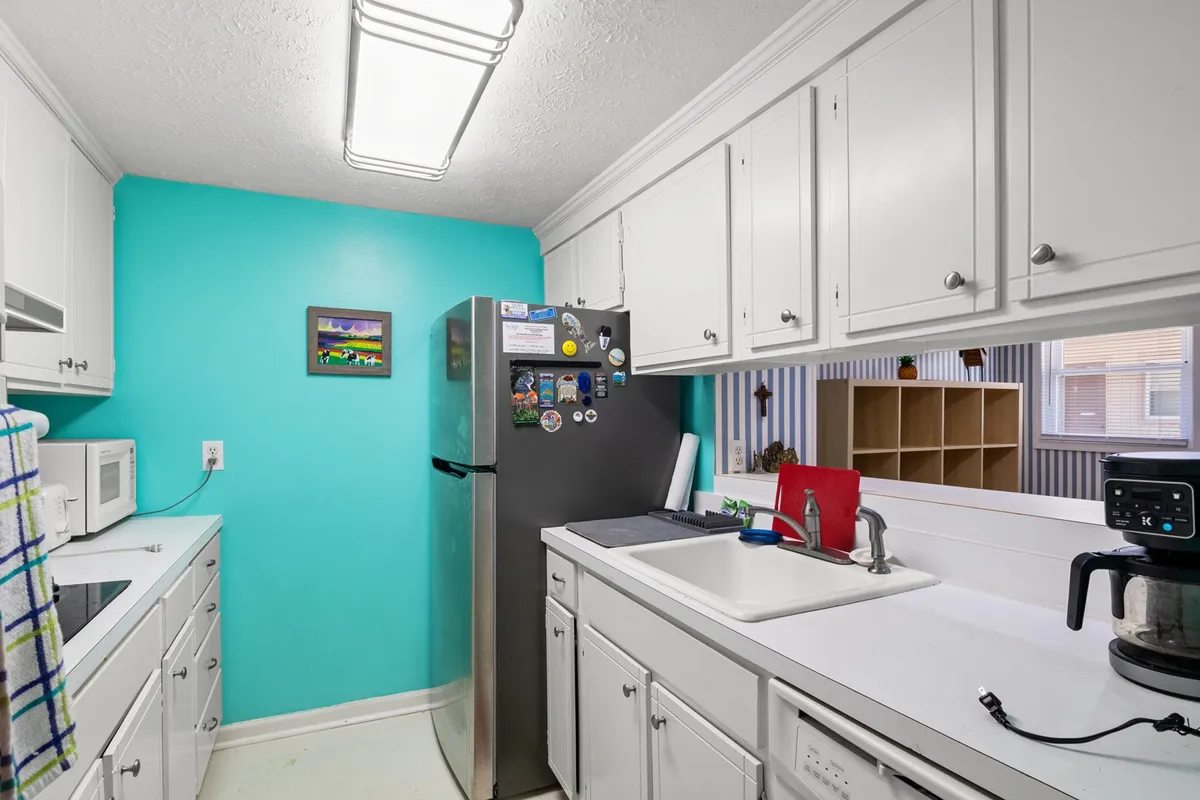 A playful, lively kitchen with a turquoise accent wall, stainless steel refrigerator, crisp white cabinets, and a convenient coffee maker. The space is designed for both functional cooking and friendly socializing, radiating a fresh, modern feel.