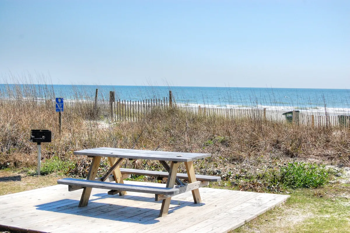 A peaceful wooden picnic table overlooks the ocean, with beach grass and a rustic fence framing the view. The calm waters and clear horizon suggest the perfect setting for morning coffee, an intimate sunset picnic, or quiet conversation. The natural elements and open-air seating evoke relaxation and privacy.
