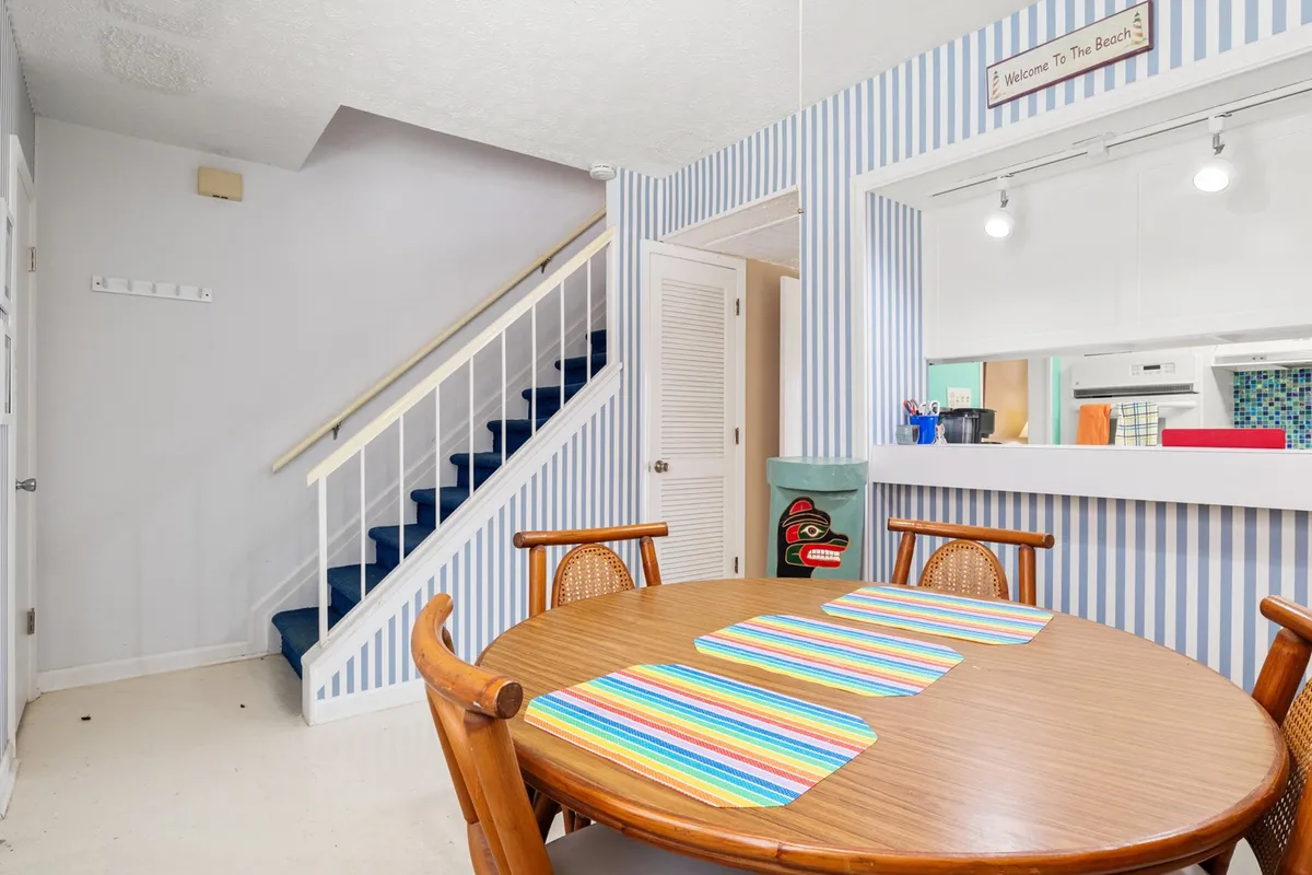 This image showcases a cozy and inviting dining area featuring a central dining table, a staircase with a striped accent wall, and a kitchen pass-through. The setting is bright and well-maintained, suggesting a well-cared-for common area ideal for family meals, social gatherings, and everyday relaxation.