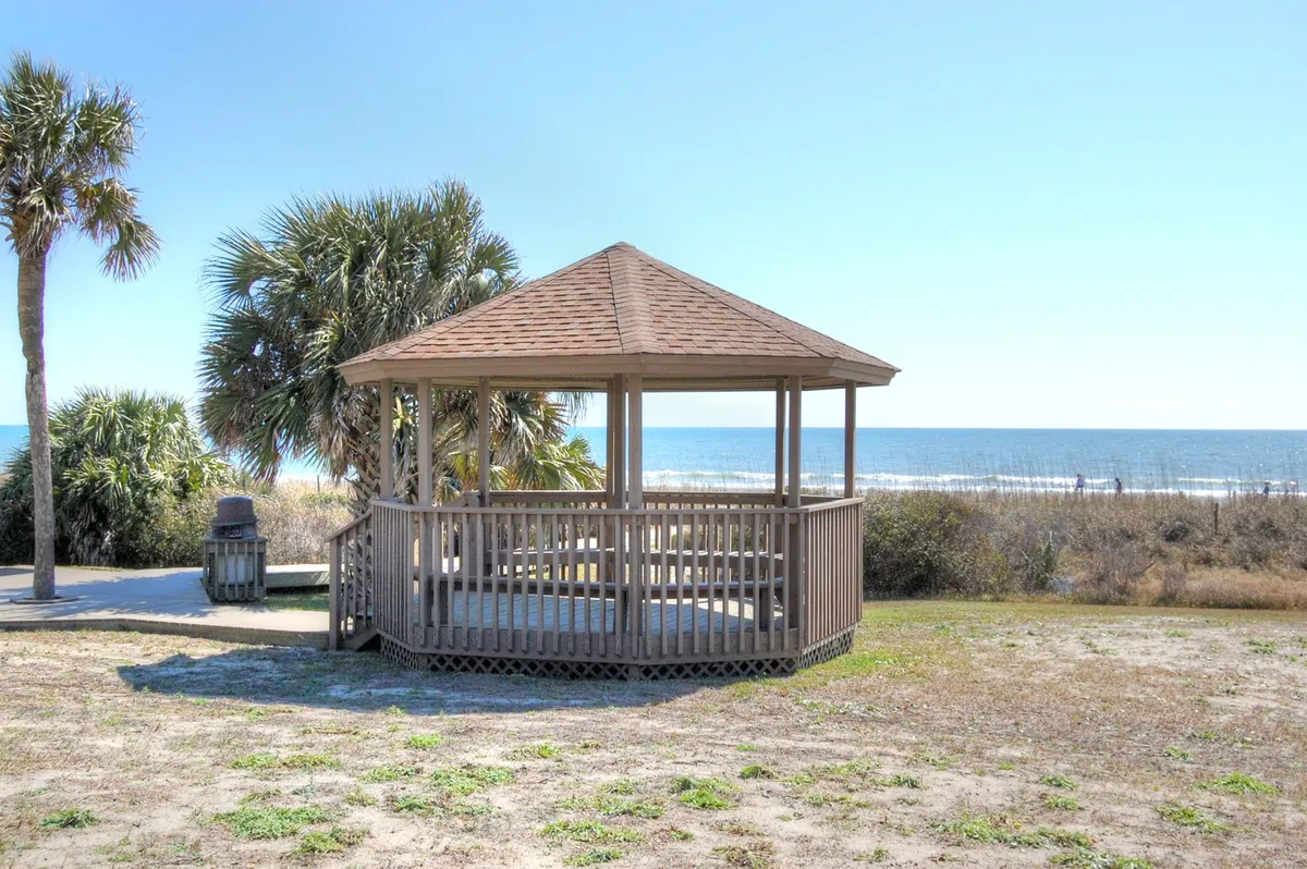 This exterior shot captures a wooden gazebo set among palm trees, with the ocean visible in the background. The space invites relaxation, picnics, and socializing under the open sky, evoking the coastal atmosphere referenced in the section.