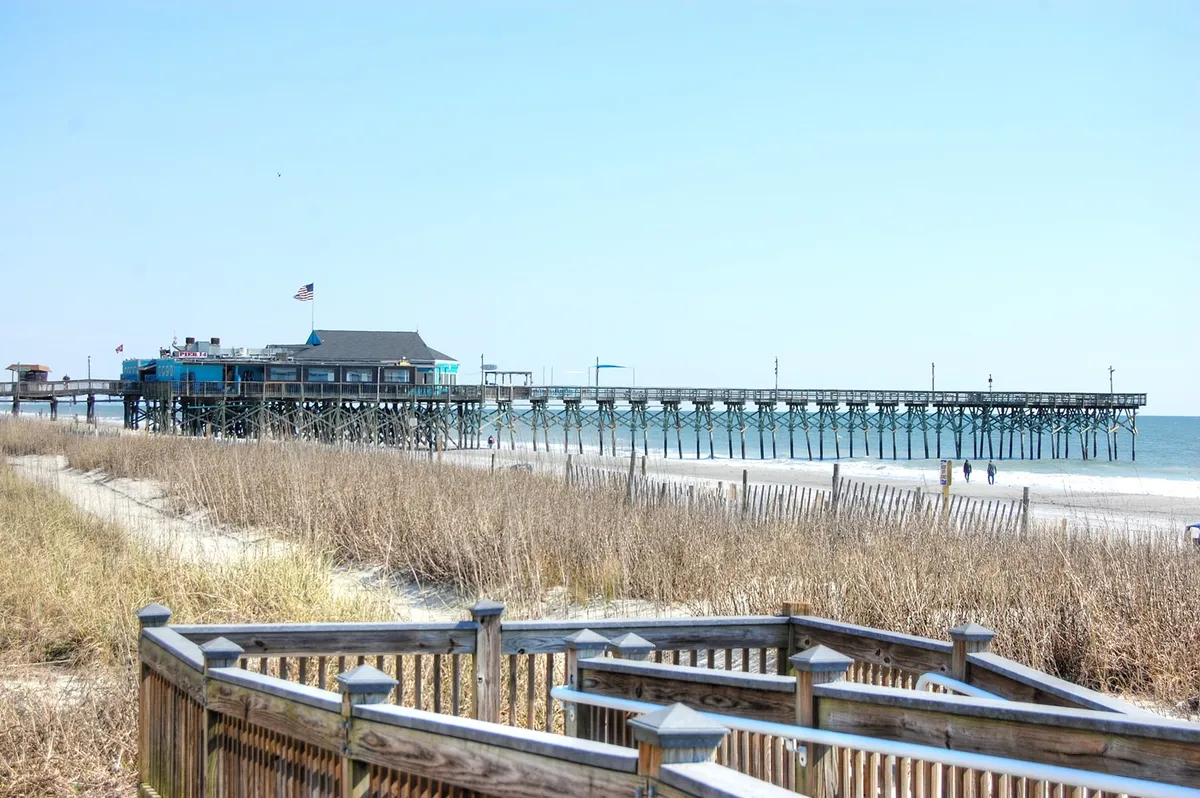 A sweeping view of the pier stretching out into the ocean, with the beach and boardwalk in the foreground and dune grass adding a natural frame. The scene includes families and friends strolling along the sand and pier, with gentle waves creating a serene, inviting atmosphere.