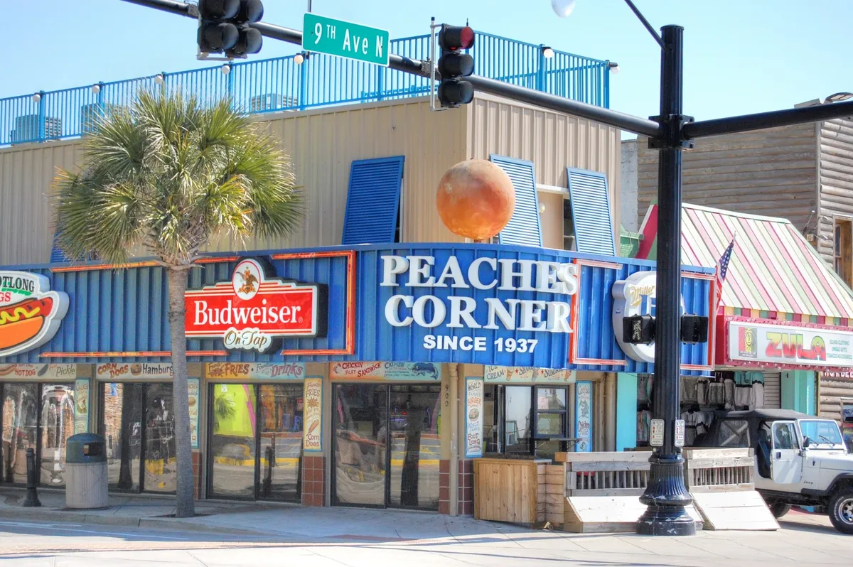 Vibrant, colorful building facades line a lively street under palm trees, with signs for Peaches Corner and Budweiser visible. People dine and socialize in the inviting atmosphere, capturing the local flavor and social scene. The setting feels casual, friendly, and quintessentially Myrtle Beach.