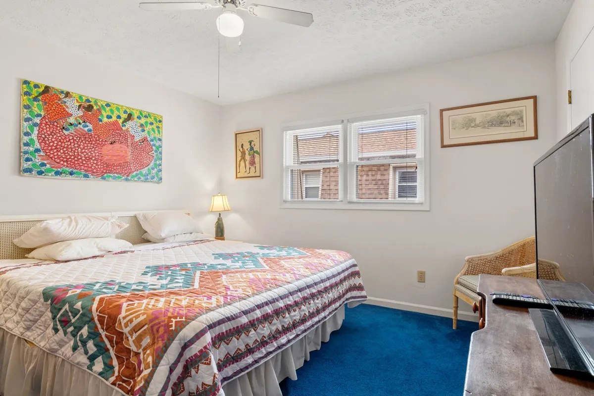 A cheerful bedroom featuring a quilted bedspread, blue carpet, colorful wall art, a nightstand with a lamp, and a TV atop a dresser. The space feels bright, inviting, and offers a cozy environment for relaxation, reading, or watching TV.