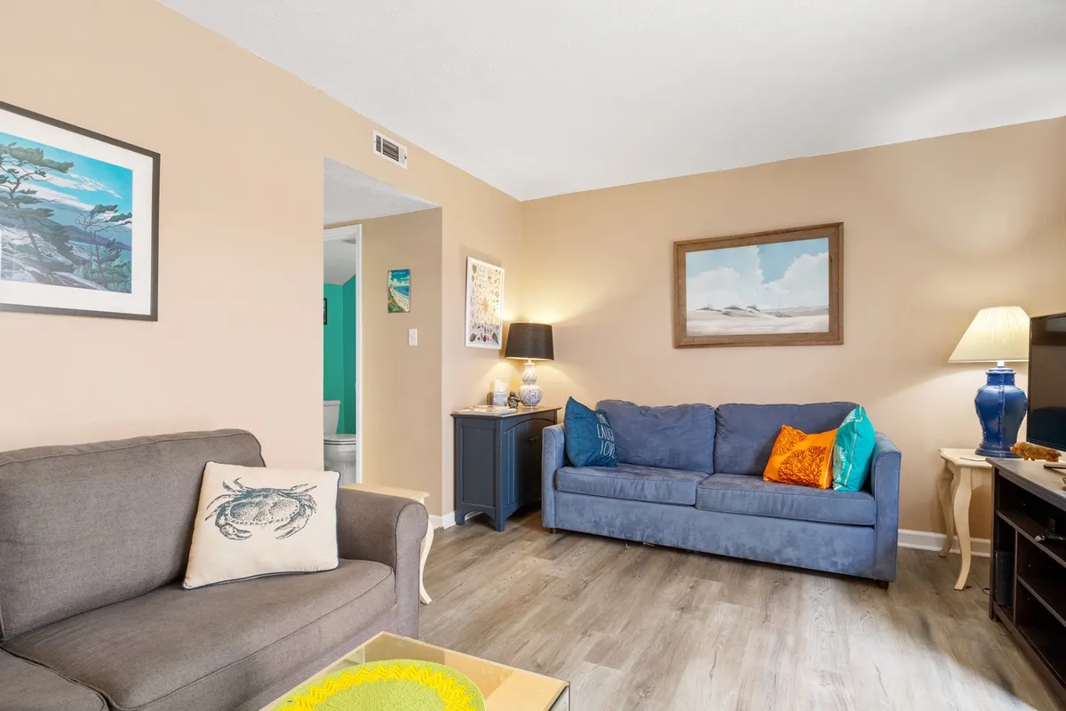 A bright and spacious living room featuring a plush blue couch adorned with decorative throw pillows. Framed artwork and a side table with a lamp add personal, welcoming touches. Natural light fills the room, highlighting the inviting atmosphere.