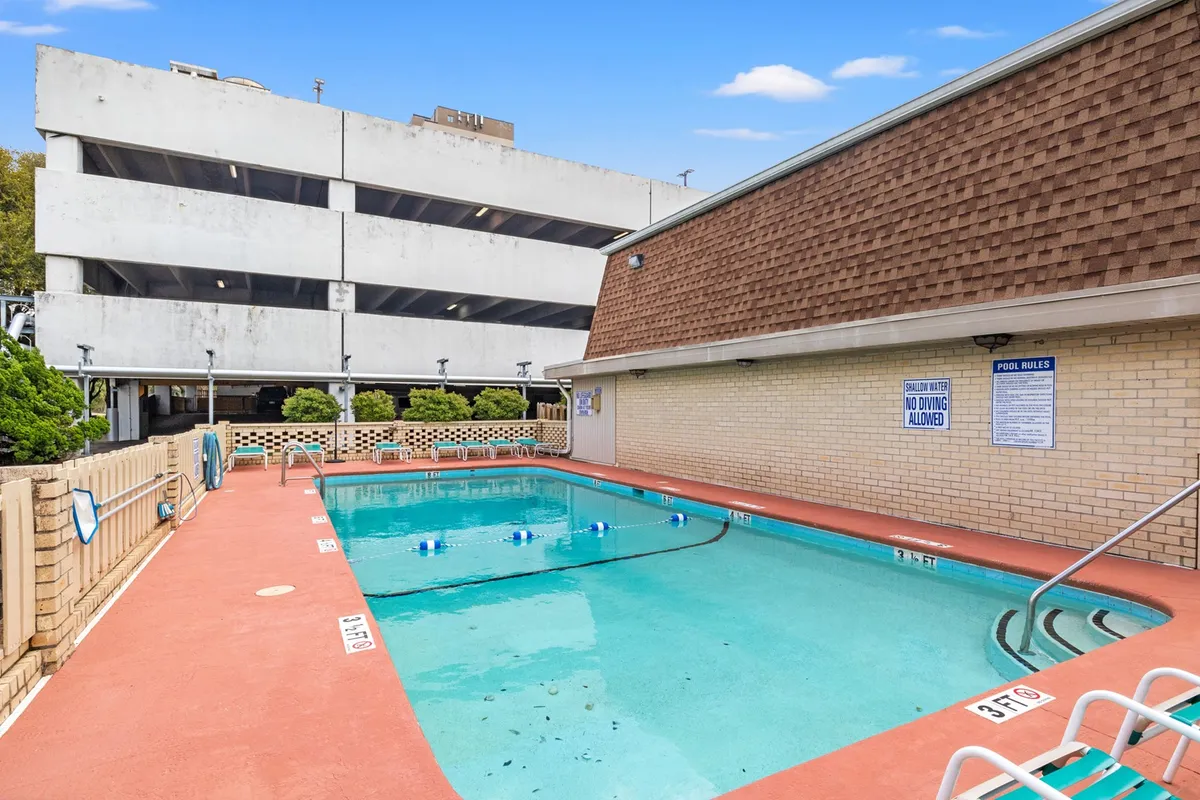 A sunlit exterior image of a swimming pool and poolside area, bordered by lush greenery and a nearby parking structure, inviting guests to swim, sunbathe, and socialize.
