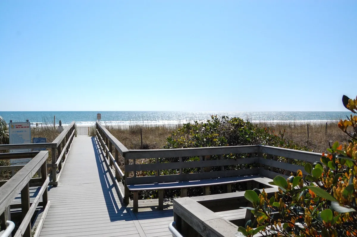 A wooden boardwalk leads through dune vegetation toward the expansive ocean, with gentle waves in the background. The scene is serene and inviting, suggesting leisurely walks, beachcombing, or simply soaking up the coastal ambiance. The open path and horizon symbolize both exploration and relaxation.