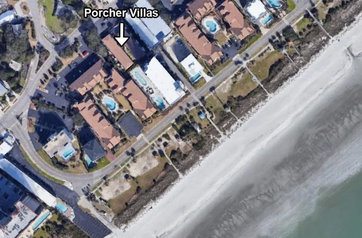 A wide-angle view of the Porcher Villas and neighboring residential buildings, with the beach visible nearby. The scene suggests easy access to both the accommodations and the shoreline, highlighting the proximity between vacation rentals and the ocean.