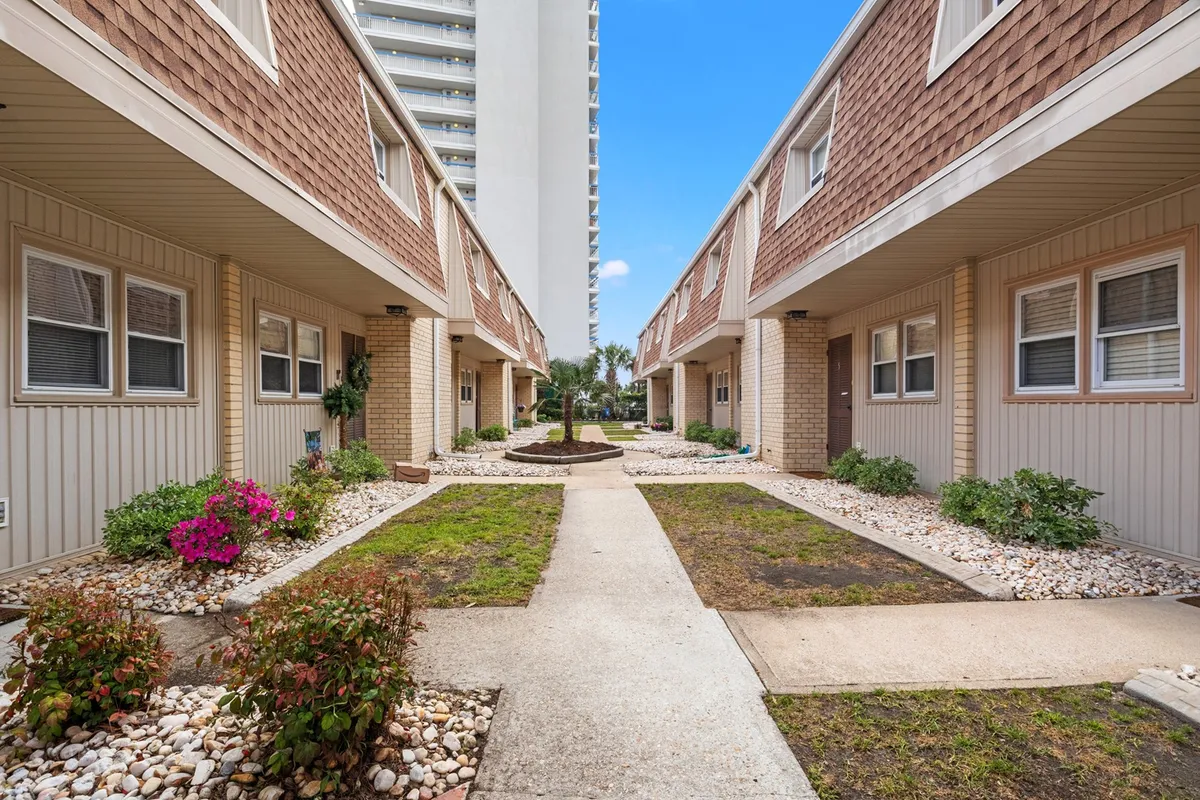 A landscaped common area with flower beds, mature trees, and attractive building facades along a sidewalk, creating a welcoming environment. The scene suggests opportunities for socializing, relaxing, and enjoying well-kept grounds.