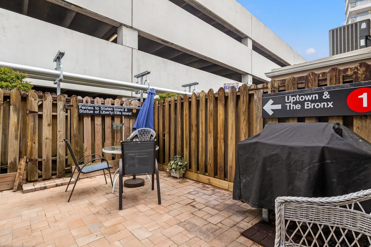 The image showcases a private back patio with inviting patio furniture, a gas grill, and a fenced area adorned with directional signs. The scene is set for casual outdoor dining and barbecuing, reflecting a relaxed, welcoming mood.