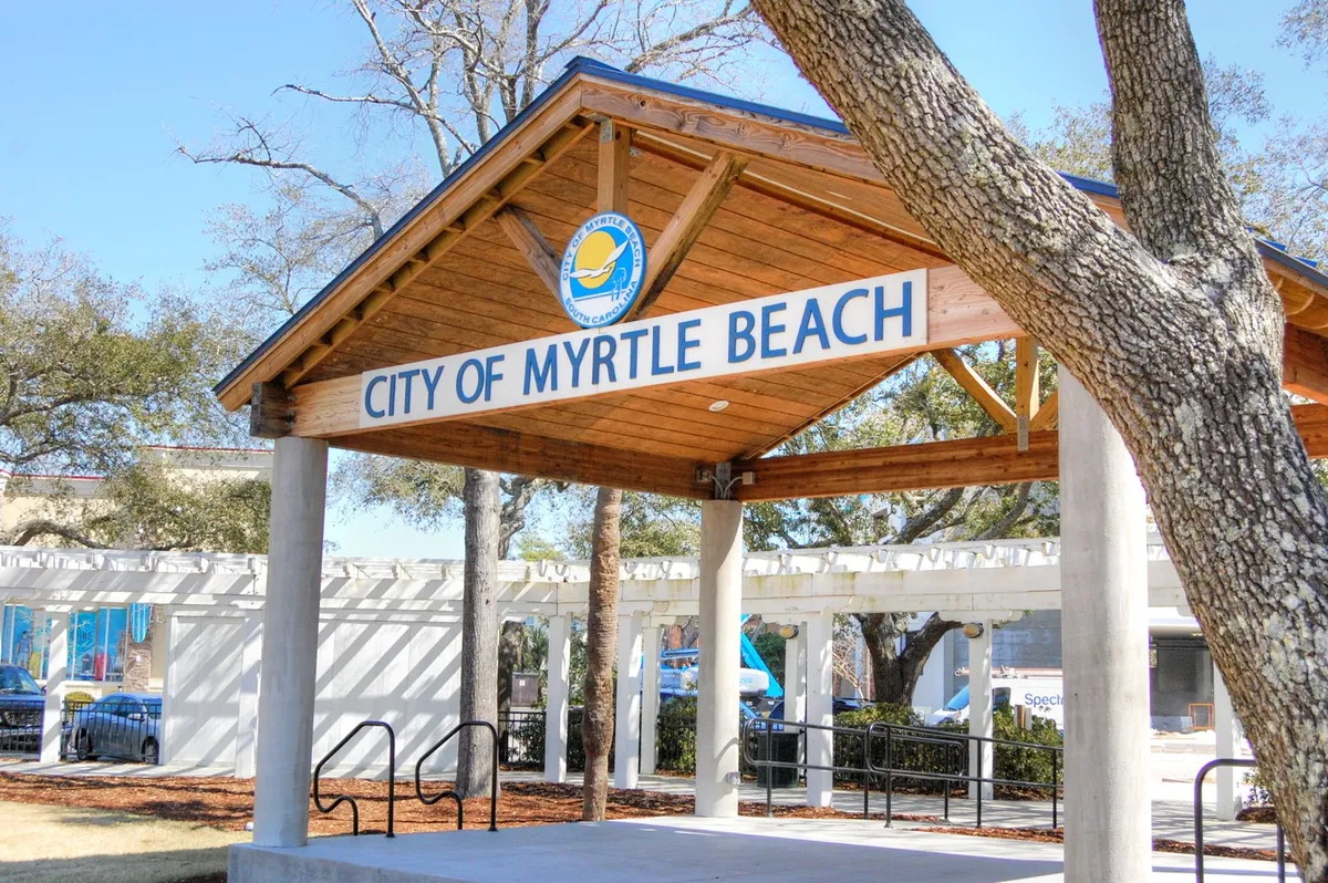The iconic 'City of Myrtle Beach' signage stands amid greenery and a wooden structure, creating a welcoming gateway vibe. The area around the sign hints at community gatherings and local happenings, reflecting the lively events and accessible local culture described in the content.