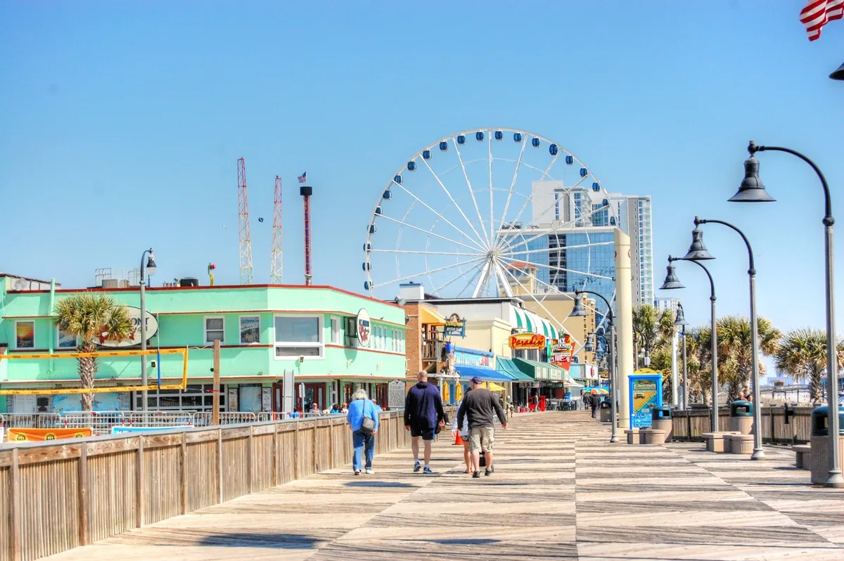 This lively exterior image captures the iconic Myrtle Beach Boardwalk, featuring the Ferris wheel, colorful buildings, and palm trees bustling with visitors walking, shopping, dining, and enjoying rides.